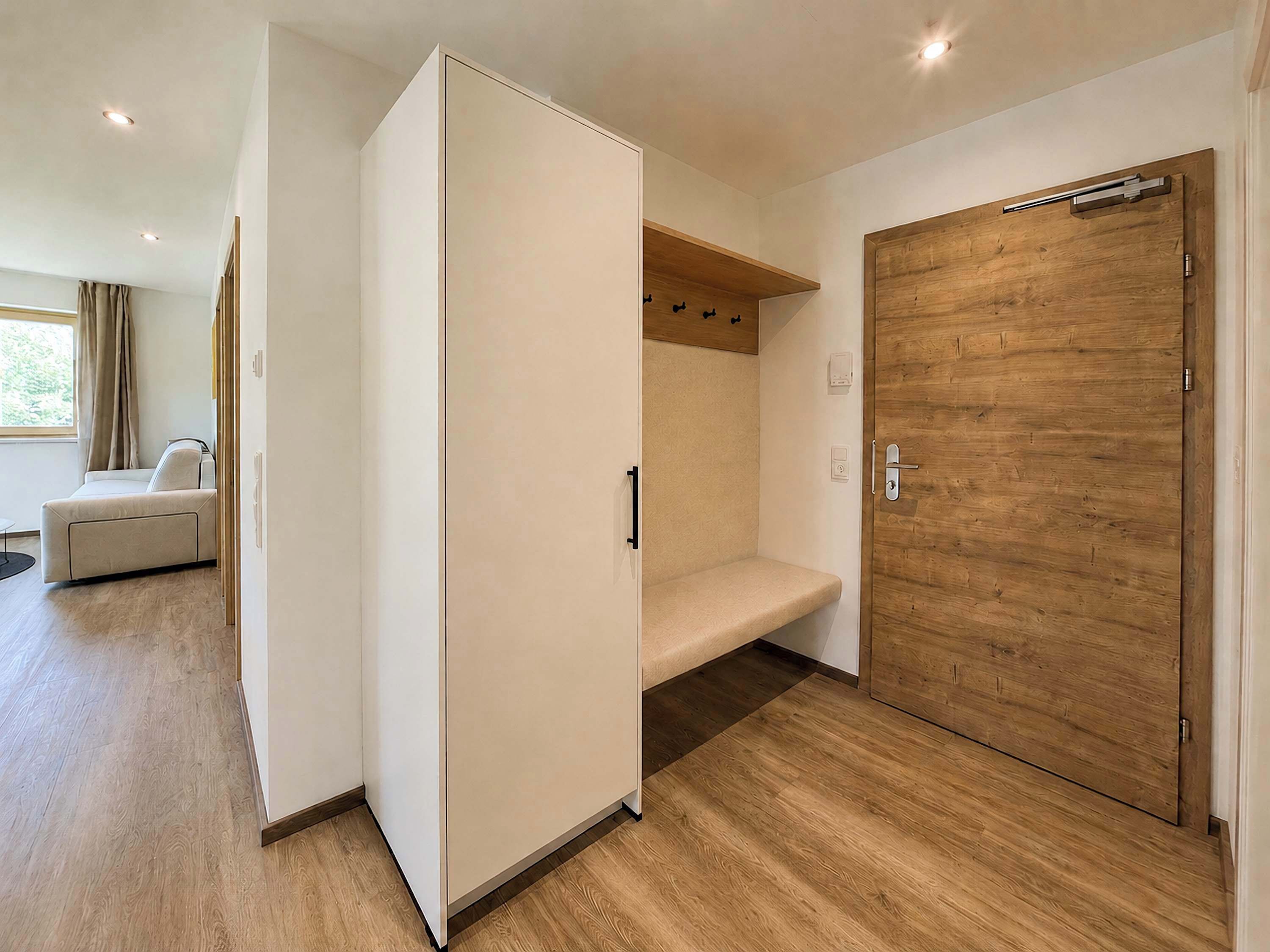 Ein modernes Zimmer mit Holztür, weißem Kleiderschrank, einer Bank und einem Fenster mit Vorhängen.