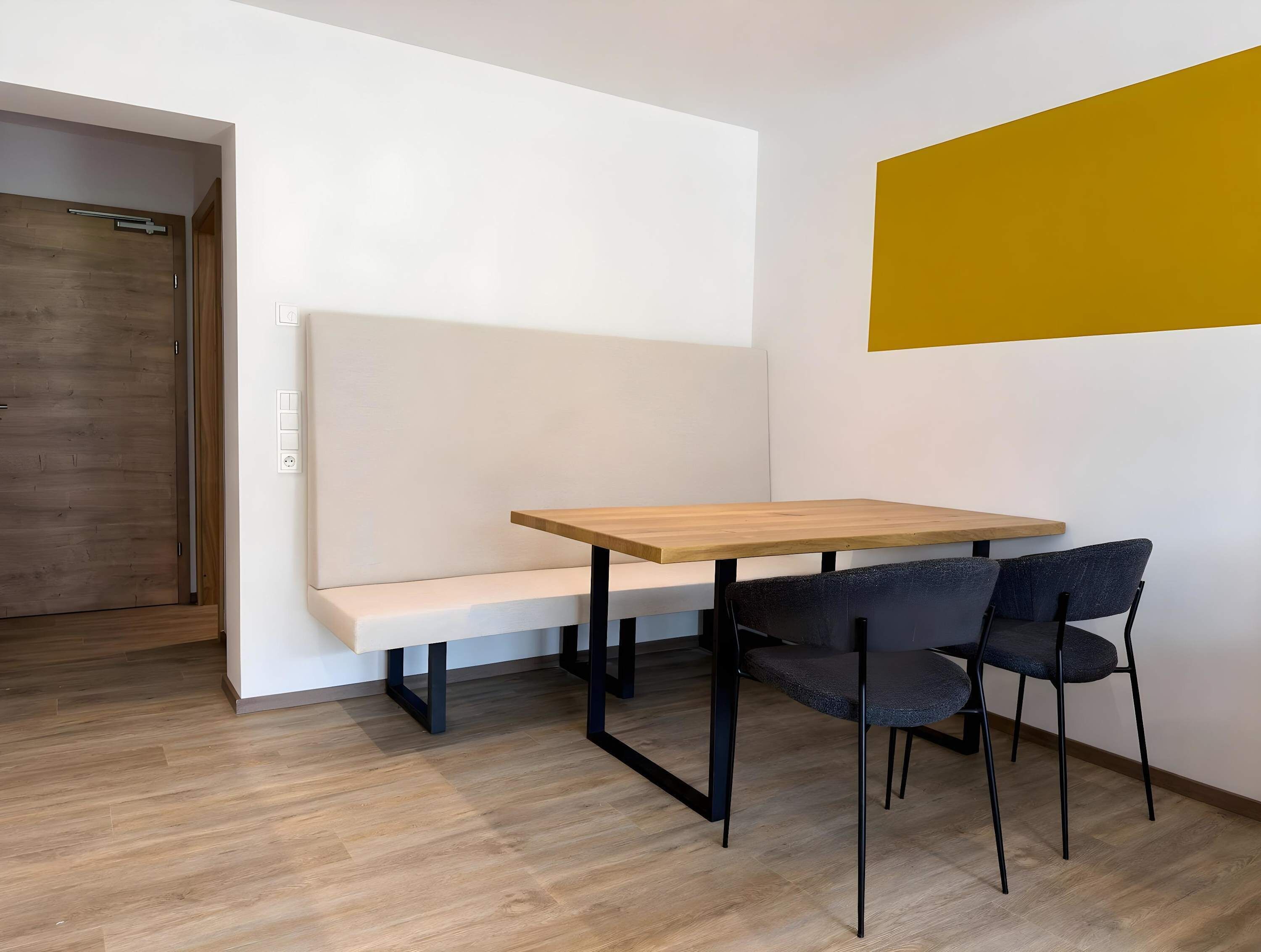 Ein kleiner, moderner Raum mit Holzboden, einer weißen Bank, einem Holztisch, zwei Stühlen und einer Tür.