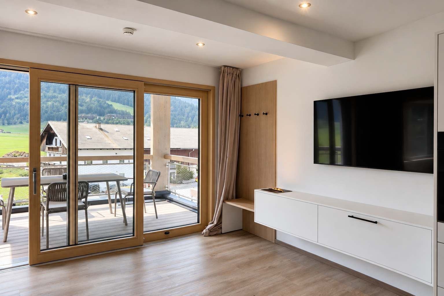 A modern interior with a sliding glass door leading to a balcony, a flat-screen TV mounted on the wall, and a minimalist wooden bench.
