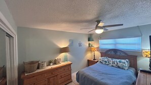Een slaapkamer met een bed, een dressoir, nachtkastjes, lampen, een plafondventilator en een raam met zonwering.