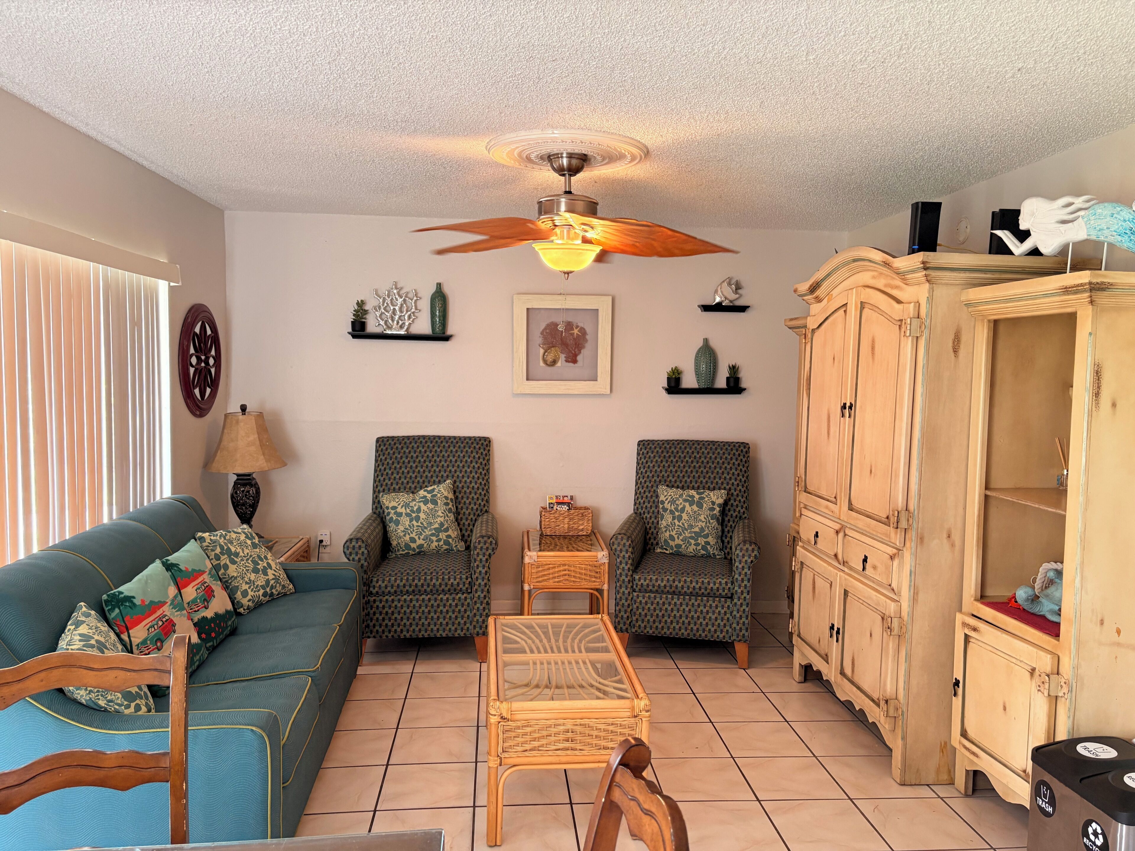 A living room with a sofa, two patterned armchairs, a coffee table, a ceiling fan, a cabinet, and a trash bin.