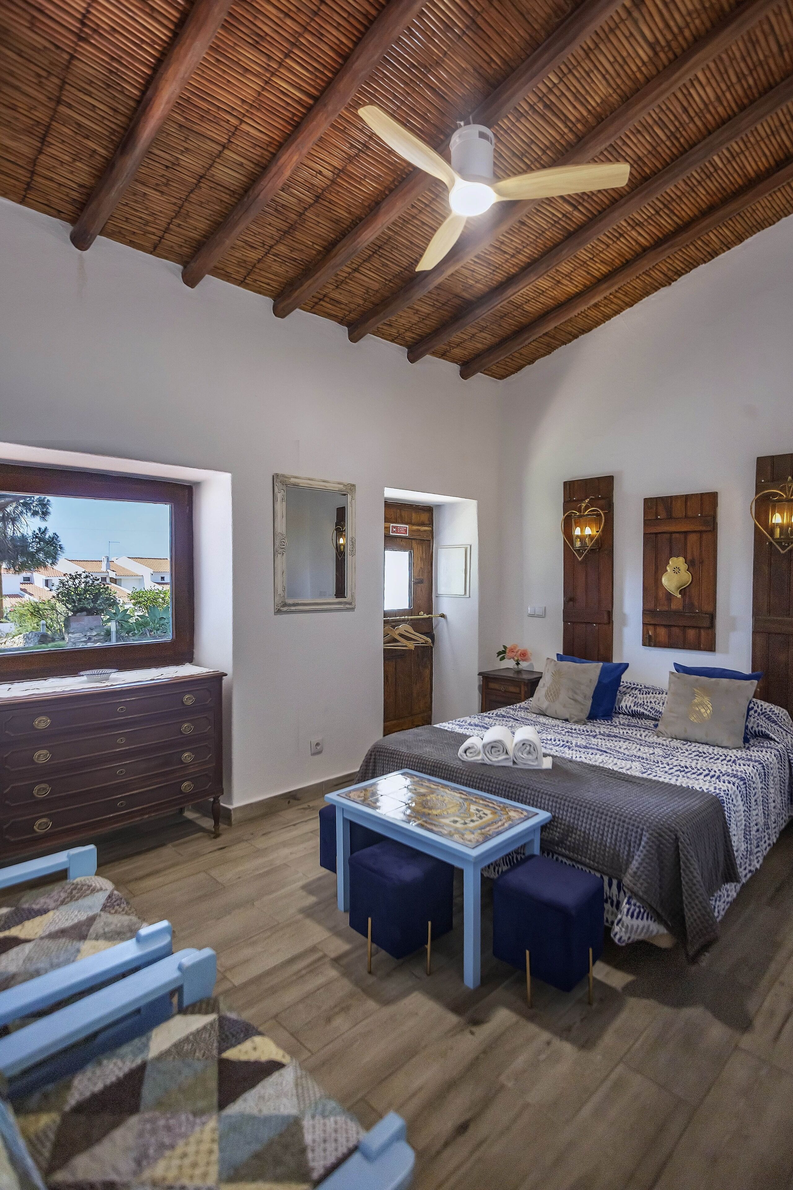 A bedroom with a bed, a desk, a chair, a ceiling fan, and a window with a view.