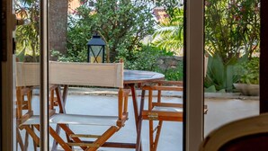 A view through a glass door to an outdoor seating area with wooden tables and chairs, a lantern, and lush greenery. - Apartment with a terrace and a view of the garden (Albufeira)
