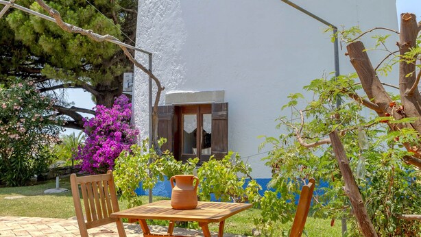 A white building with blue trim, a patio with wooden furniture, and a tree with purple flowers. - Historic Windmill – Panoramic Views (Albufeira)
