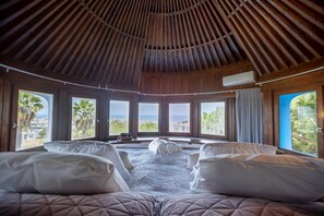 Quarto com teto de madeira, janelas amplas, duas camas com almofadas e vista para o mar.