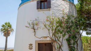 A cylindrical building with a blue and white exterior, a wooden door, and a small garden area with plants and a tree. - Historic Windmill – Panoramic Views (Albufeira)