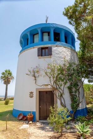 Um edifício cilíndrico com exterior azul e branco, uma porta de madeira e uma pequena área de jardim com plantas e uma árvore.