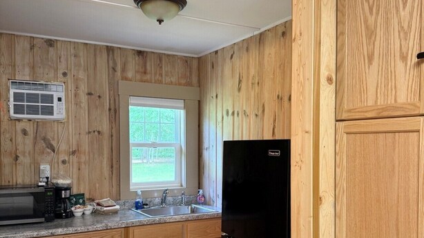 Eine Küche mit Holzschränken, einem schwarzen Kühlschrank, einer Mikrowelle und einem Fenster mit Blick auf Grünflächen.