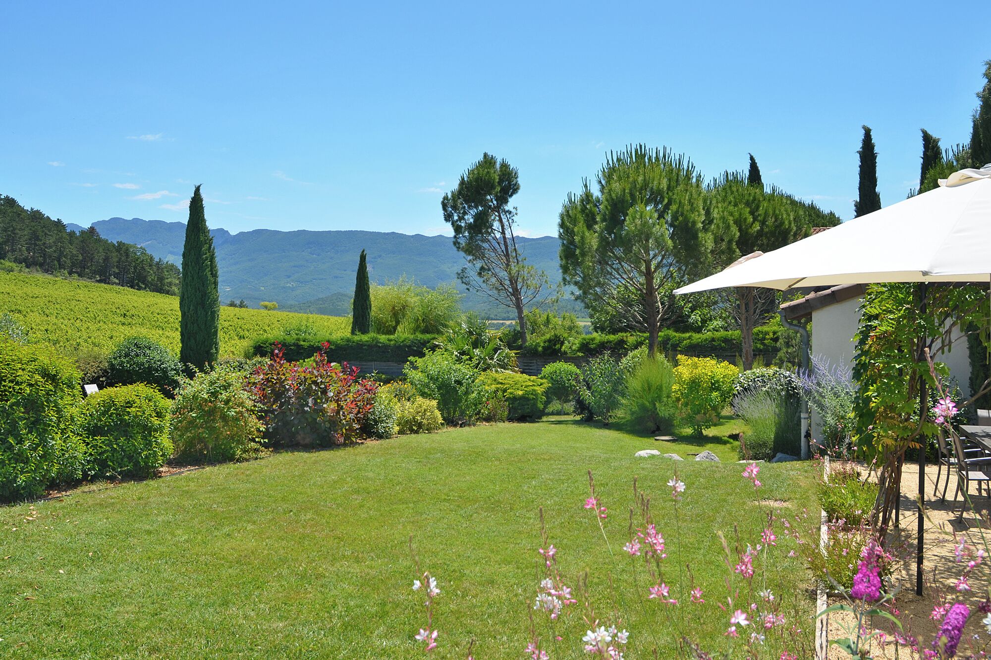 A well-maintained garden with a lawn, various shrubs, and trees, a patio area with outdoor furniture, and a view of distant mountains.