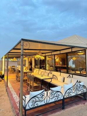 An outdoor seating area with tables and chairs, a covered section with a glass wall, and a decorative metal bench. - Mama Desert Camp (Merzouga)