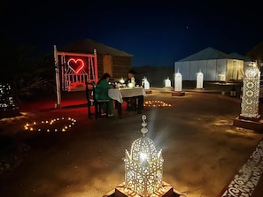 An outdoor dining area with tables and chairs, surrounded by decorative lanterns and a heart-shaped light installation. - Mama Desert Camp (Merzouga)