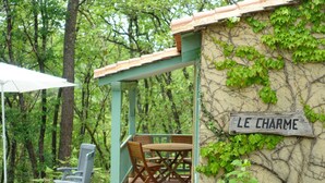 Een overdekt terras met een tafel en stoelen, een bord met de tekst 'LE CHARME' en een stenen muur begroeid met groen.