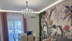 Ein modernes Wohnzimmer mit Sofa, Sesseln, einer Chandelüster, Wandmalerei und einem Essbereich.