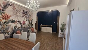 A dining area with a wooden table, white chairs, a sofa, and a wall with tropical plant murals.