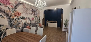 A dining area with a wooden table, white chairs, a sofa, and a wall with tropical plant murals.
