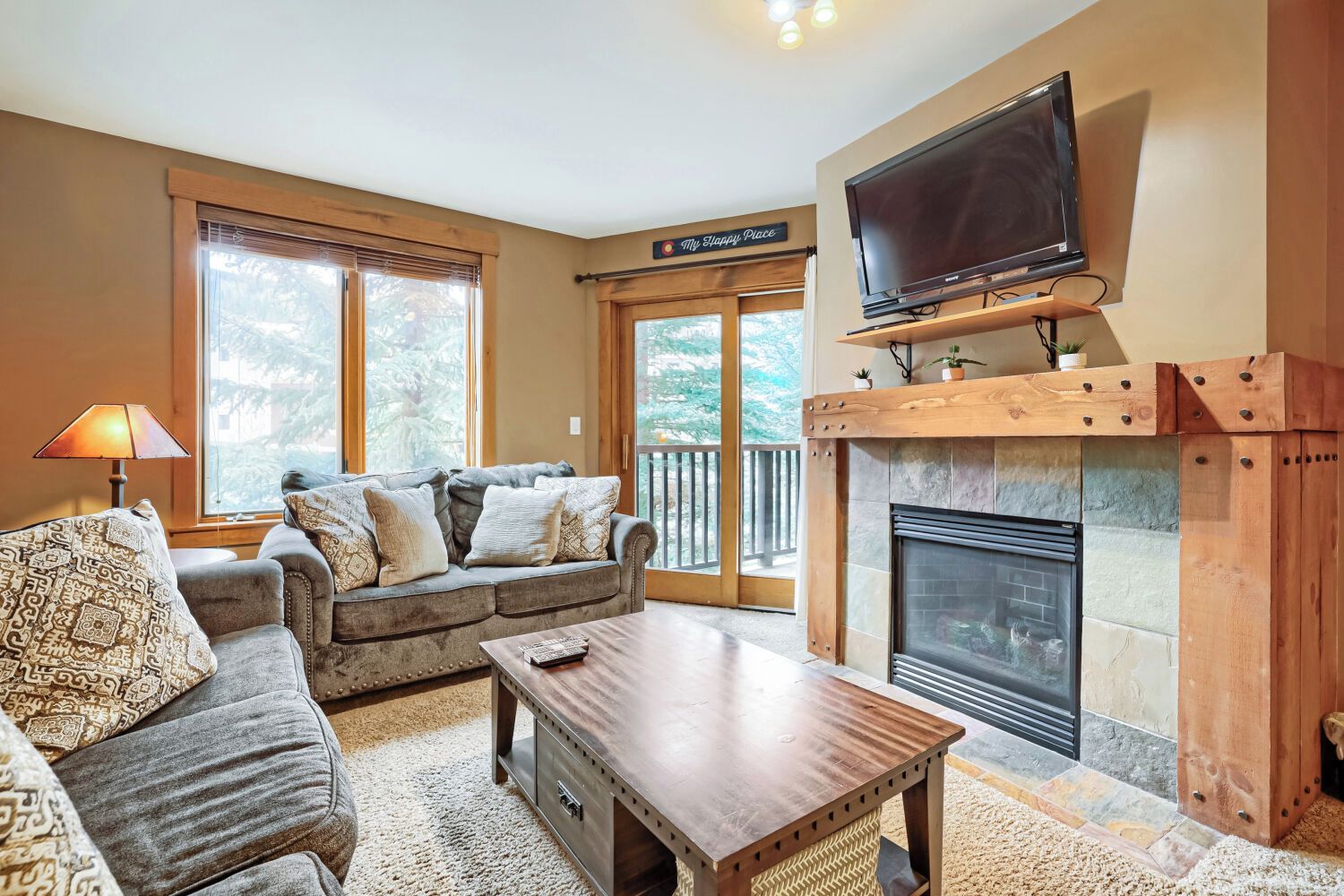 A living room with a fireplace, a television mounted on a wooden beam above it, and a couch with patterned cushions.