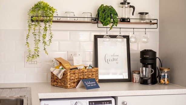 A kitchen with a coffee maker, a framed welcome sign, and a basket of items on the counter. - 3-Bed House Sleeps 6 with Driveway Parking · 10 Min to Knutsford (Cheshire West and Chester)