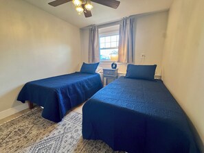 Ein Zimmer mit zwei Betten, einem Deckenventilator, einem Fenster mit Vorhängen und einem Nachttisch mit Lampe.