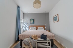 Ein Schlafzimmer mit Bett, Nachttisch, Pendelleuchte, Fenster mit Vorhängen und einem Wandbild.