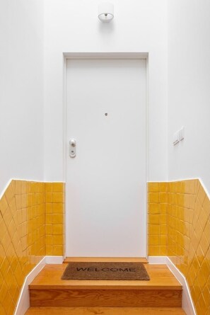 Eine weiße Tür mit Knauf und kleinem Sprechloch, neben gelb gefliesten Wänden, die zu einer Holztreppe mit Fußmatte führt.