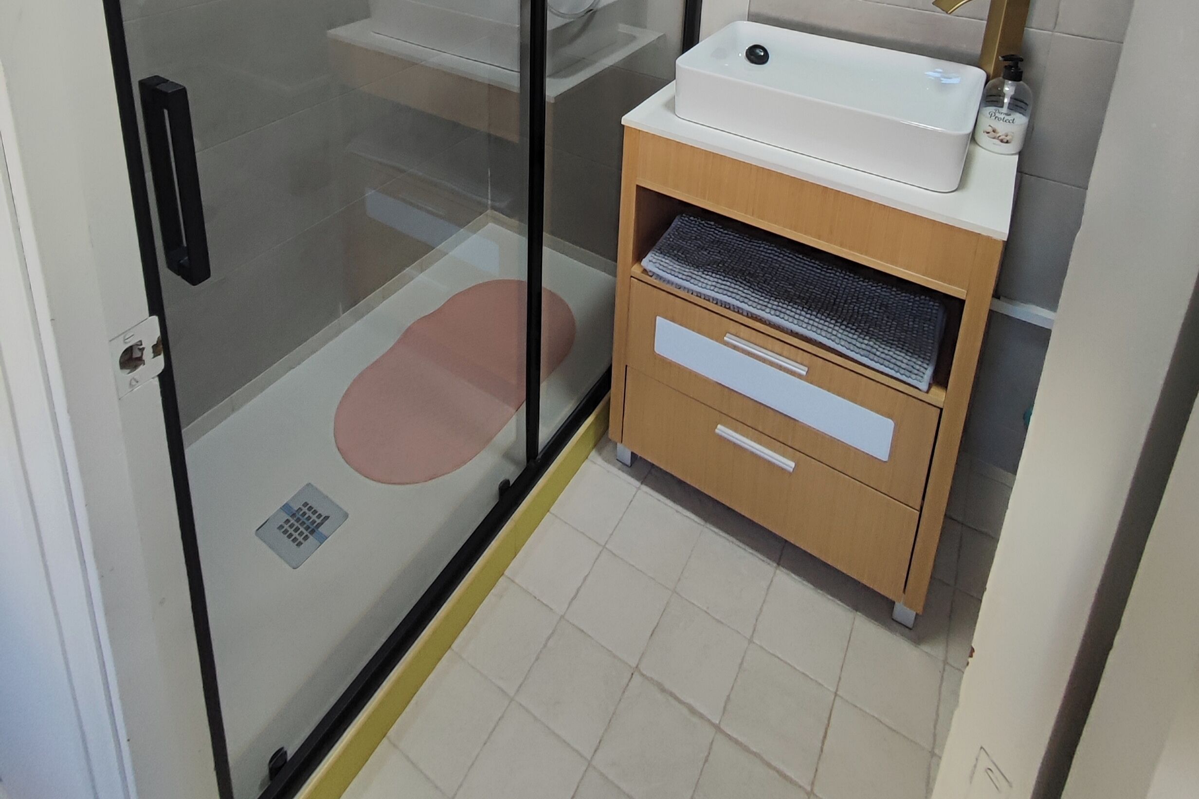 Ein Badezimmer mit Glastrennwand, einem Waschbecken, einem Holzkabinent mit Schubladen und einem gefliesten Boden.