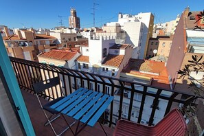 Un balcón con mesa y sillas, con vista a un paisaje urbano que muestra diversos edificios y tejados.