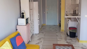 Una cocina compacta con microondas, refrigerador y lavadora. Se ve un sofá amarillo con un cojín de colores en primer plano.