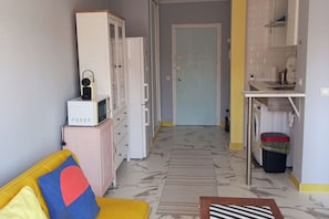 Eine kompakte Küche mit Mikrowelle, Kühlschrank und Waschmaschine. Ein gelbes Sofa mit einem bunten Kissen ist im Vordergrund zu sehen.