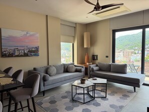 A modern living room with a sofa, coffee table, and chairs, large windows with a city view, and a ceiling fan. - Sunset Lovers Paradise w/ rooftop shared pool & jacuzzi w/ perfect location (Puerto Vallarta)