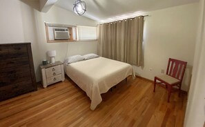 A bedroom with a bed, a nightstand, a chair, a dresser, a window with curtains, and an air conditioning unit. - Living on a budget? Look here! (Hays)