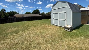 A small, single-story shed with a gabled roof and a door, situated on a grassy yard with a wooden fence in the background. - Living on a budget? Look here! (Hays)