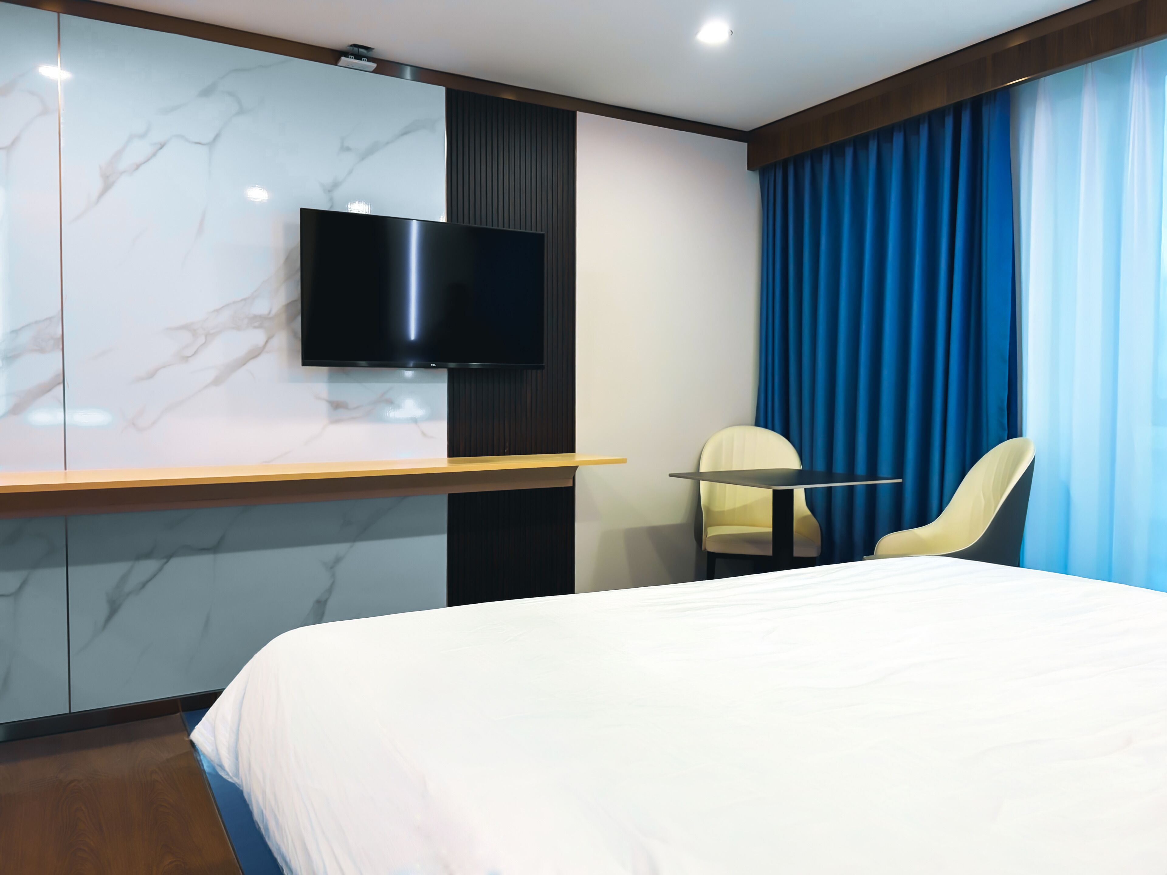 Una habitación de hotel moderna con una cama grande, un televisor de pantalla plana montado en una pared de mármol, una mesita y una silla.