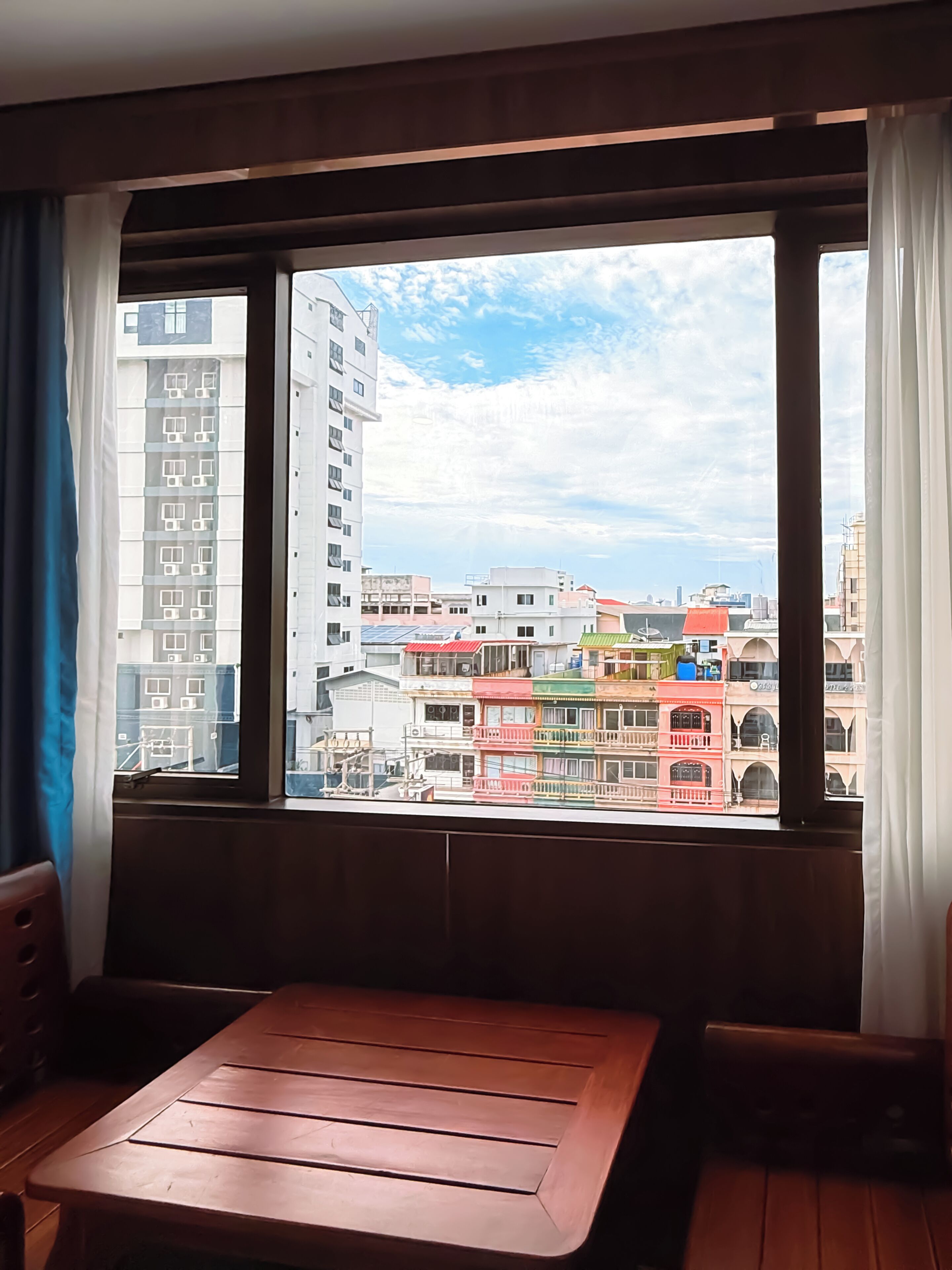 Una habitación con una mesa y sillas de madera, una ventana con cortinas y vistas a edificios coloridos.
