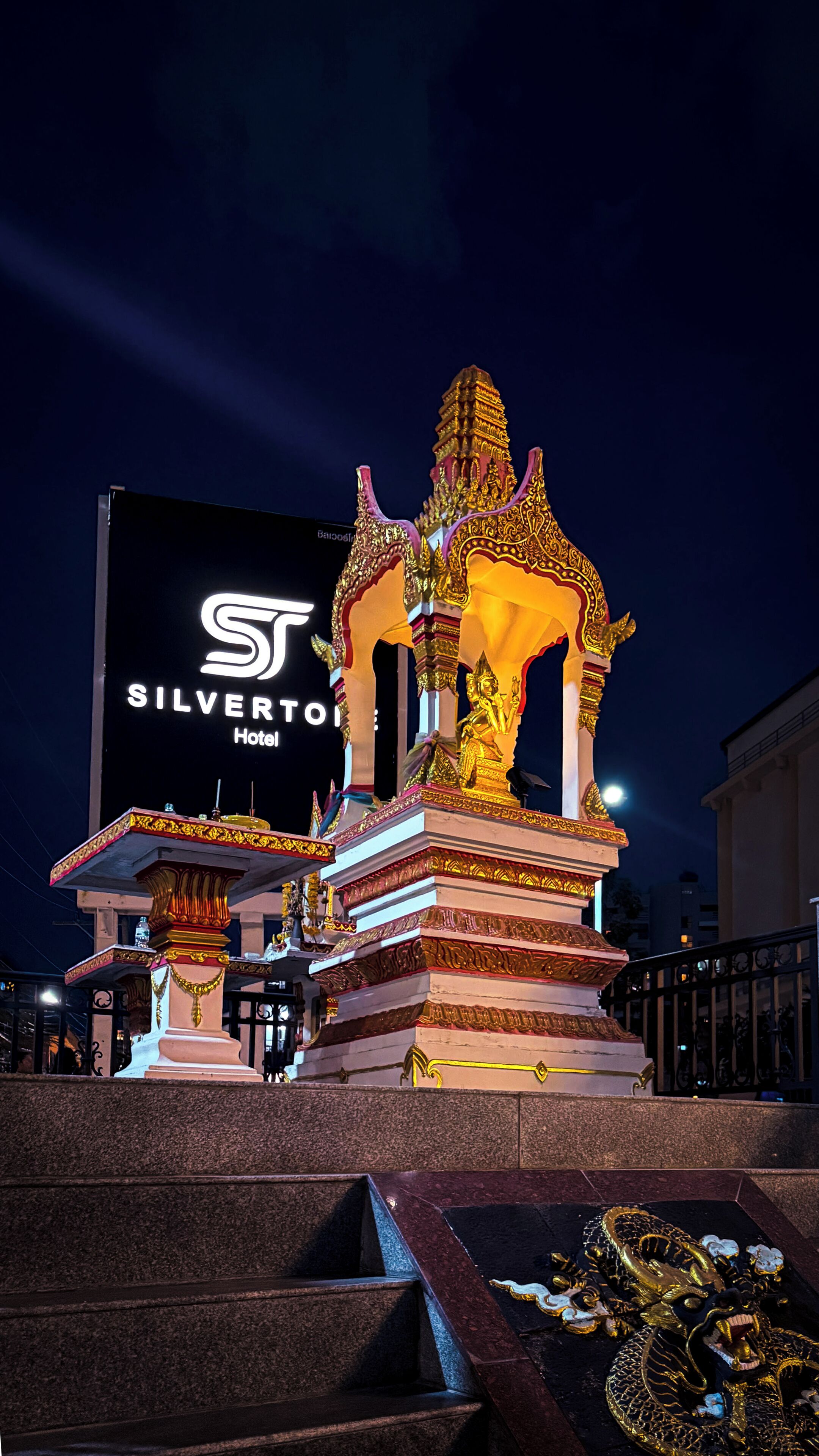 A night view of a hotel with a large illuminated sign, a golden statue, and traditional architectural elements.