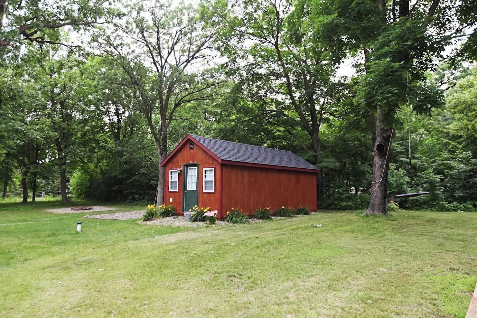 A small red wooden cabin with a green door, surrounded by a grassy area and trees.