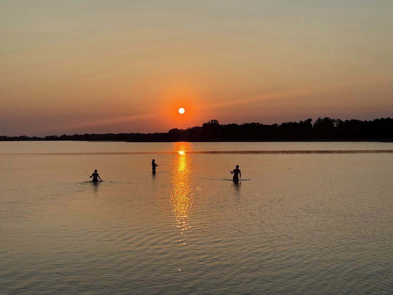 Un lac paisible au coucher du soleil, où trois personnes pêchent.