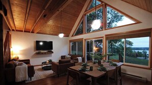 A spacious living area with a high ceiling, wooden beams, a flat-screen TV, a dining table set for four, and large windows offering a view of the outdoors. - Leaf Lake Cabin, Henning MN (Henning)
