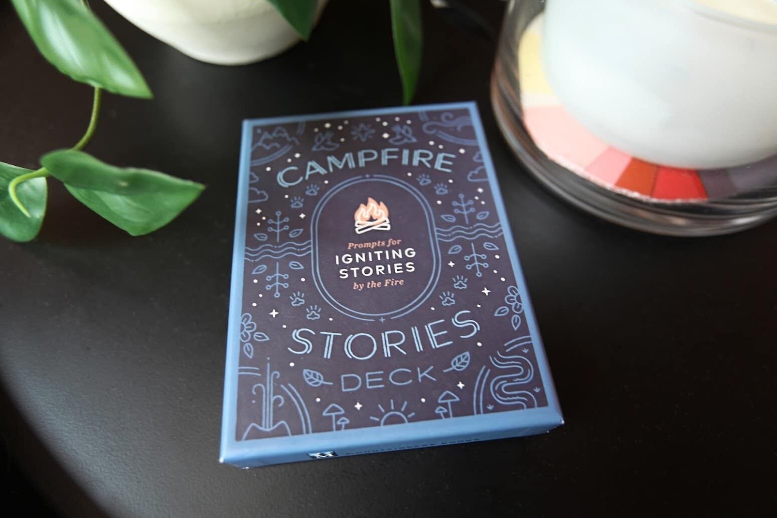 Un llibre anomenat 'Campfire Stories Deck' amb un foc de camp i una espelma al fons.