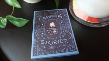 Un llibre anomenat 'Campfire Stories Deck' amb un foc de camp i una espelma al fons.