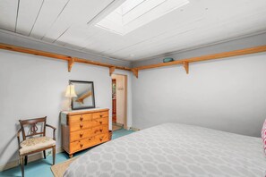 Ein Schlafzimmer mit einem Bett, einer Holzkommode, einem Sessel, einem Spiegel und einem Oberlicht.