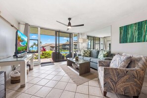 A modern living room with a glass door leading to a balcony, a ceiling fan, and a television on a glass table. - Mahina Surf 123 (Lahaina)