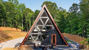 Une cabane triangulaire en bois avec une façade en verre, un espace extérieur aménagé pour s’asseoir et un chemin de gravier.