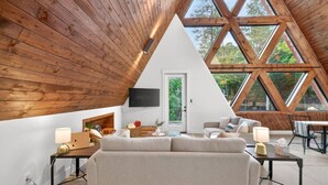 Une pièce de style « loft » avec un plafond en pente, des poutres en bois, un canapé, une télévision et de grandes fenêtres.
