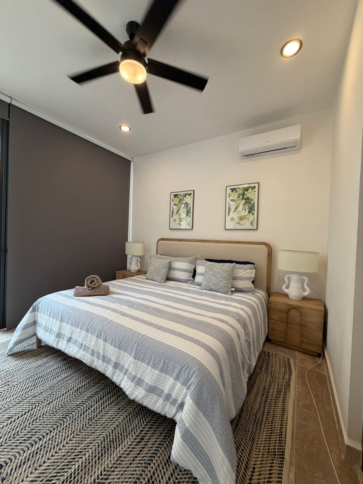 Un dormitorio con cama, ventilador de techo, aire acondicionado instalado en la pared y dos cuadros enmarcados.