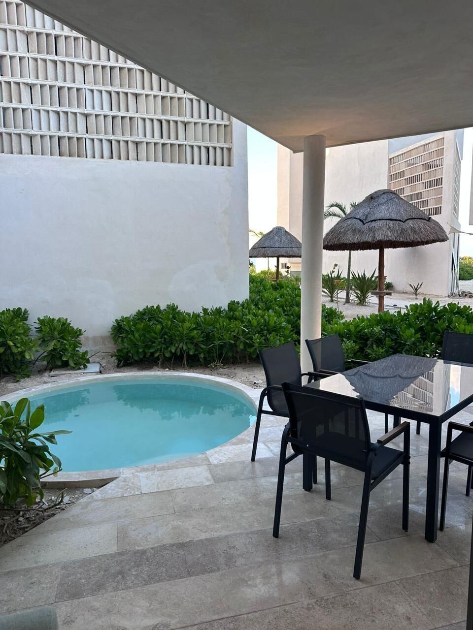 A poolside area with a table and chairs, surrounded by greenery and a building in the background.