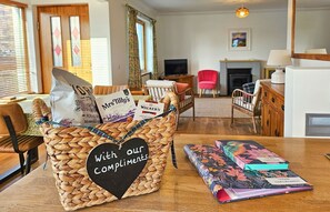 A cozy living room with a wicker basket labeled 'With Our Compliments' containing gift bags, a fireplace, and a dining area with a table and chairs. - Lochside - sleeps 6 guests  in 3 bedrooms (Dornie)