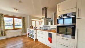 Une cuisine avec des armoires blanches, un plan de travail en bois, un four encastré et une vue sur l’extérieur grâce à de grandes fenêtres.