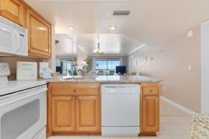 A kitchen with wooden cabinets, a white microwave, a dishwasher, and a granite countertop. - Carlos Pointe #534 Heavenly Sunsets (Fort Myers Beach)