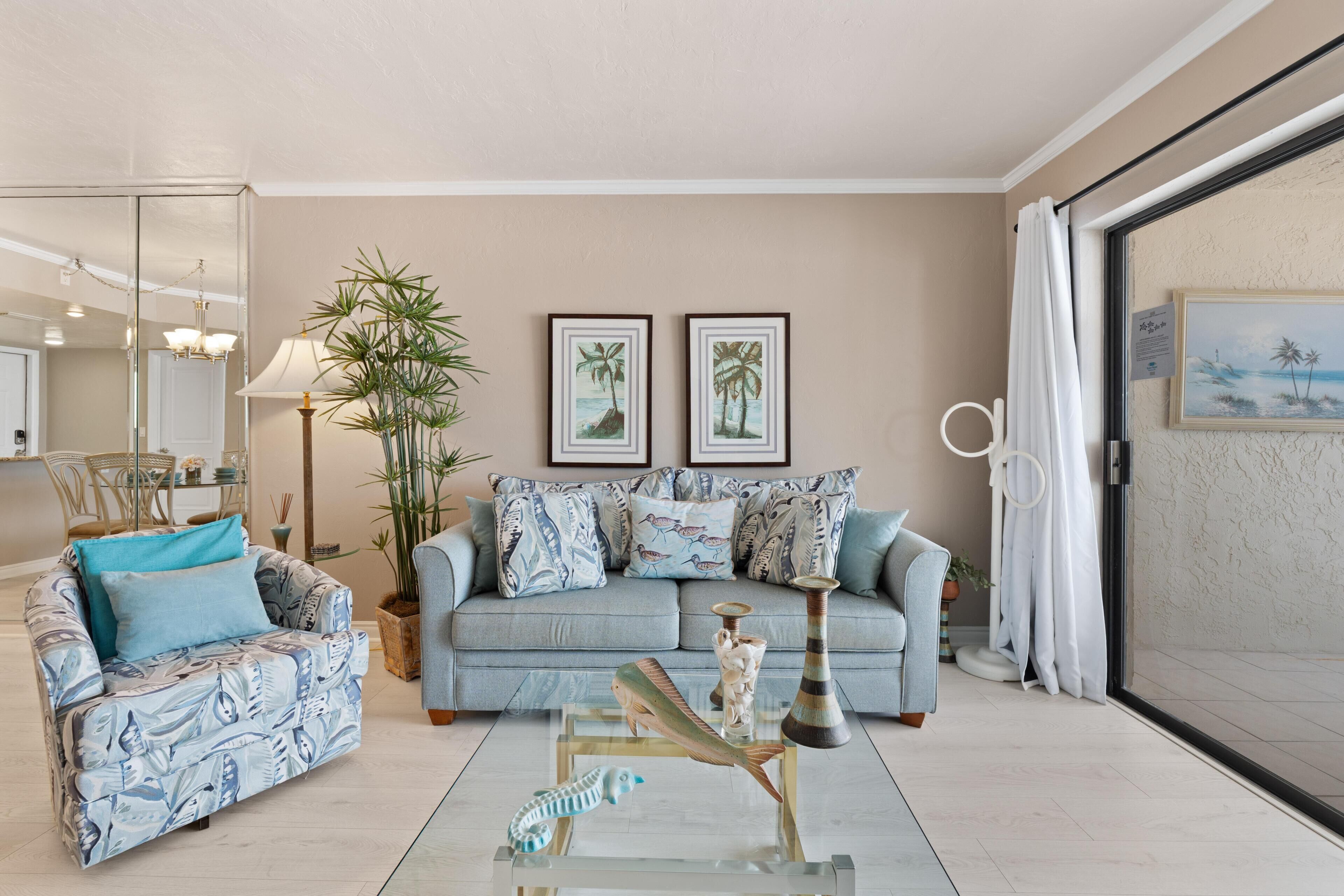 Un salon comprenant un canapé, un fauteuil et une table basse en verre. Deux tableaux encadrés ornent le mur, ainsi qu’une plante en pot et une grande fenêtre avec des rideaux.