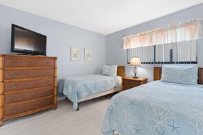 Une chambre avec un téléviseur posé sur une commode, deux lits, une table de chevet et une fenêtre avec des rideaux.